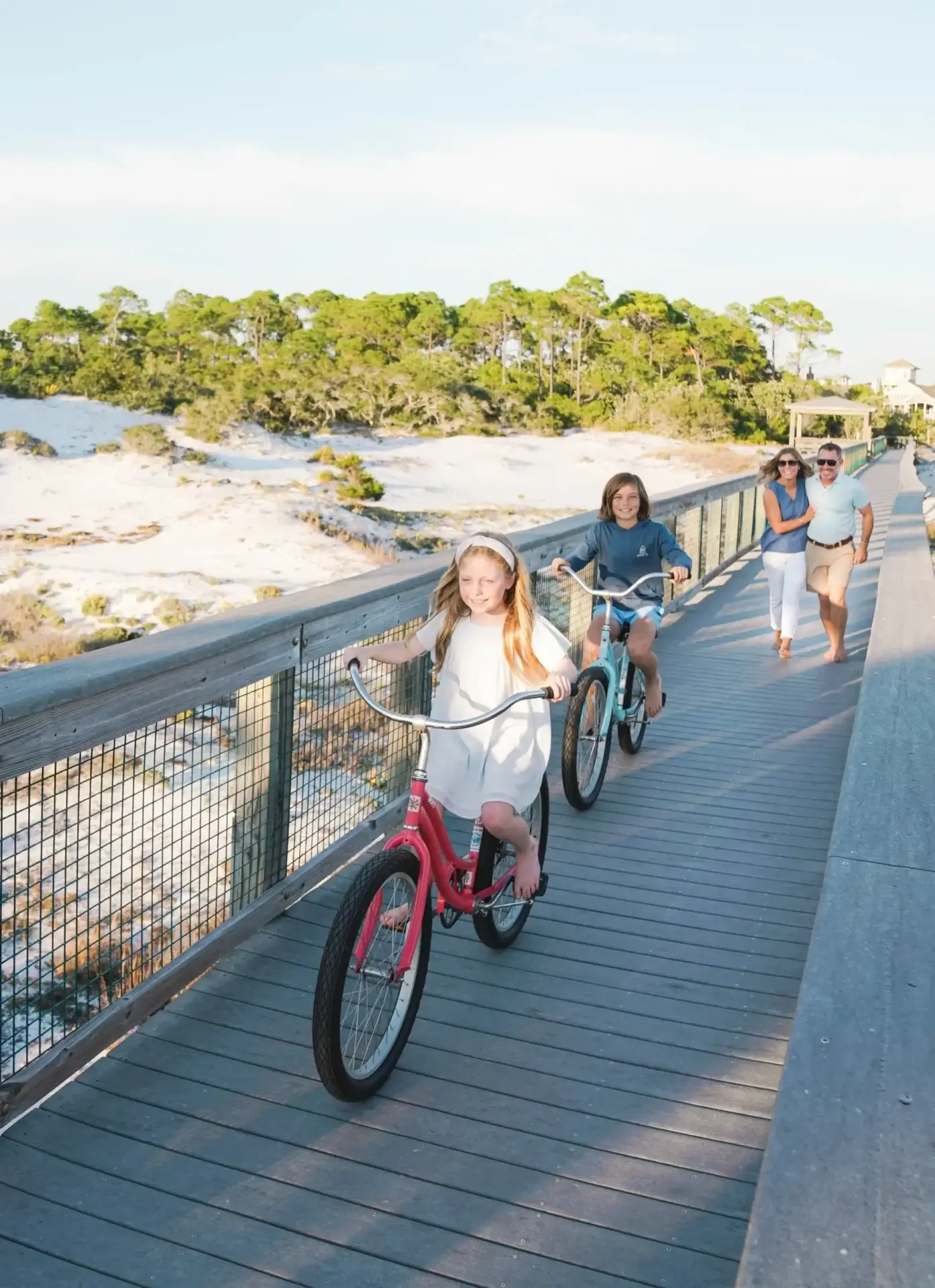 Children biking on boardwalk near beach dunes.